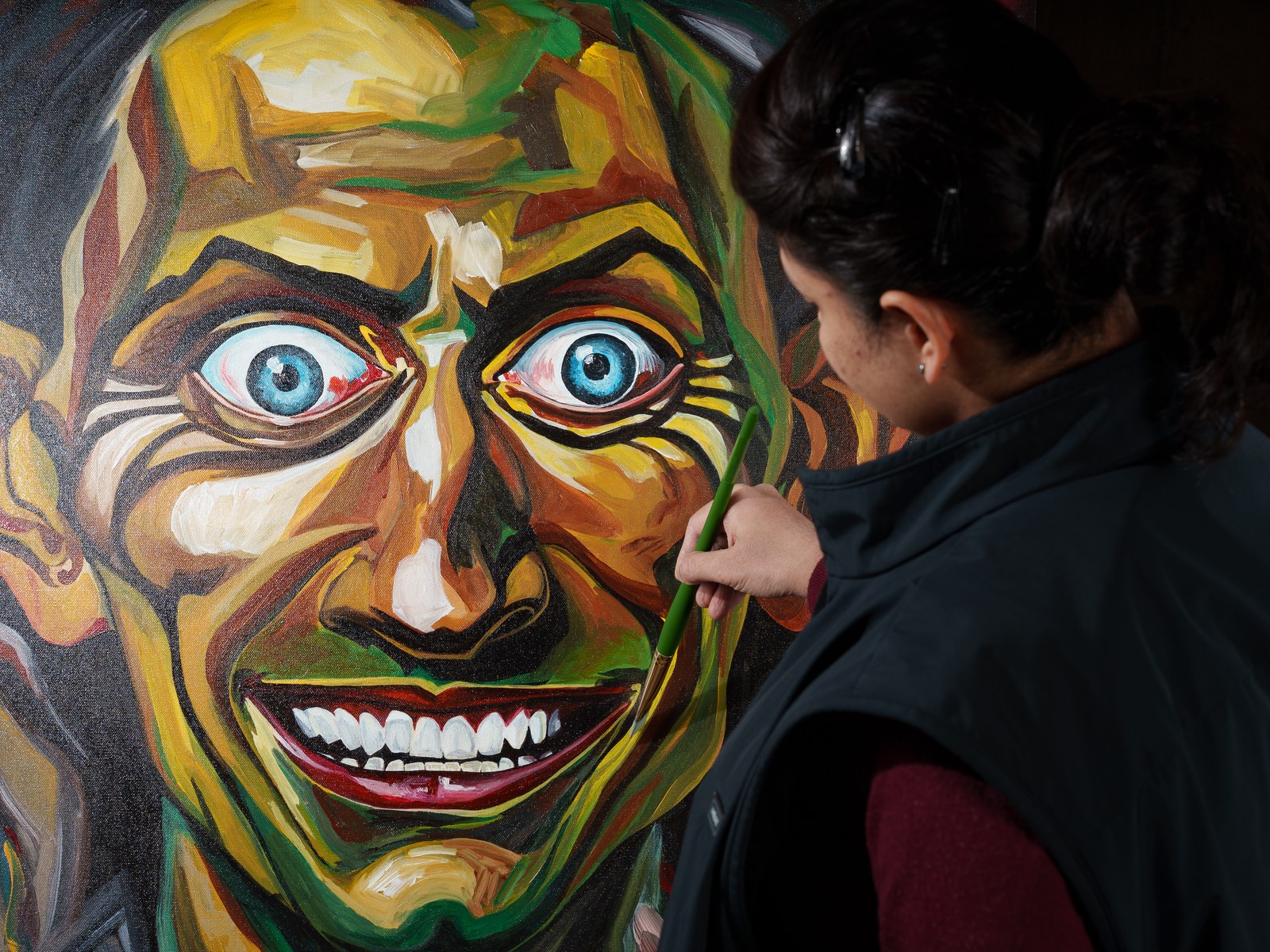 The artist faces her painting, green brush raised toward the figure's cheek. The Insane Smile Longest, 2026, by Payal Agarwal.